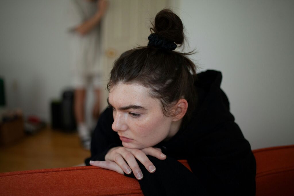 A person with dark hair in a bun leans over the back of an orange couch, looking thoughtful or concerned—perhaps reflecting on relapse prevention. The background is blurred, showing another person standing and a door.
