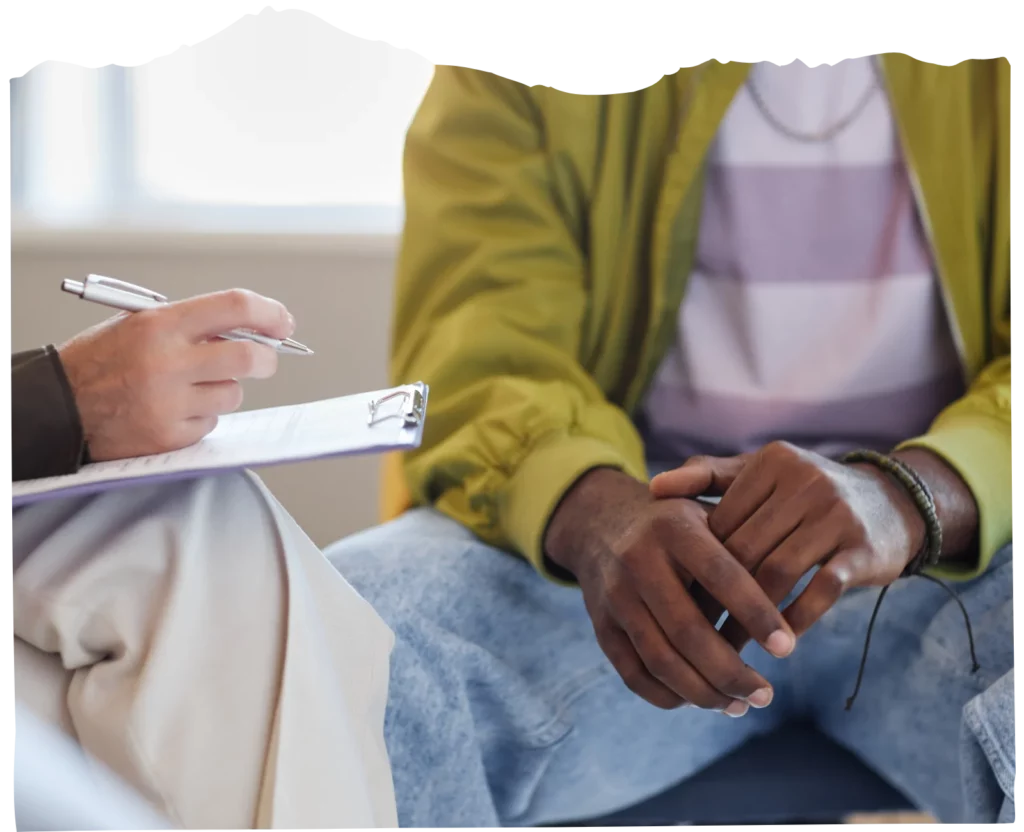 Two people seated; one is holding a clipboard and pen while the other sits with hands clasped.