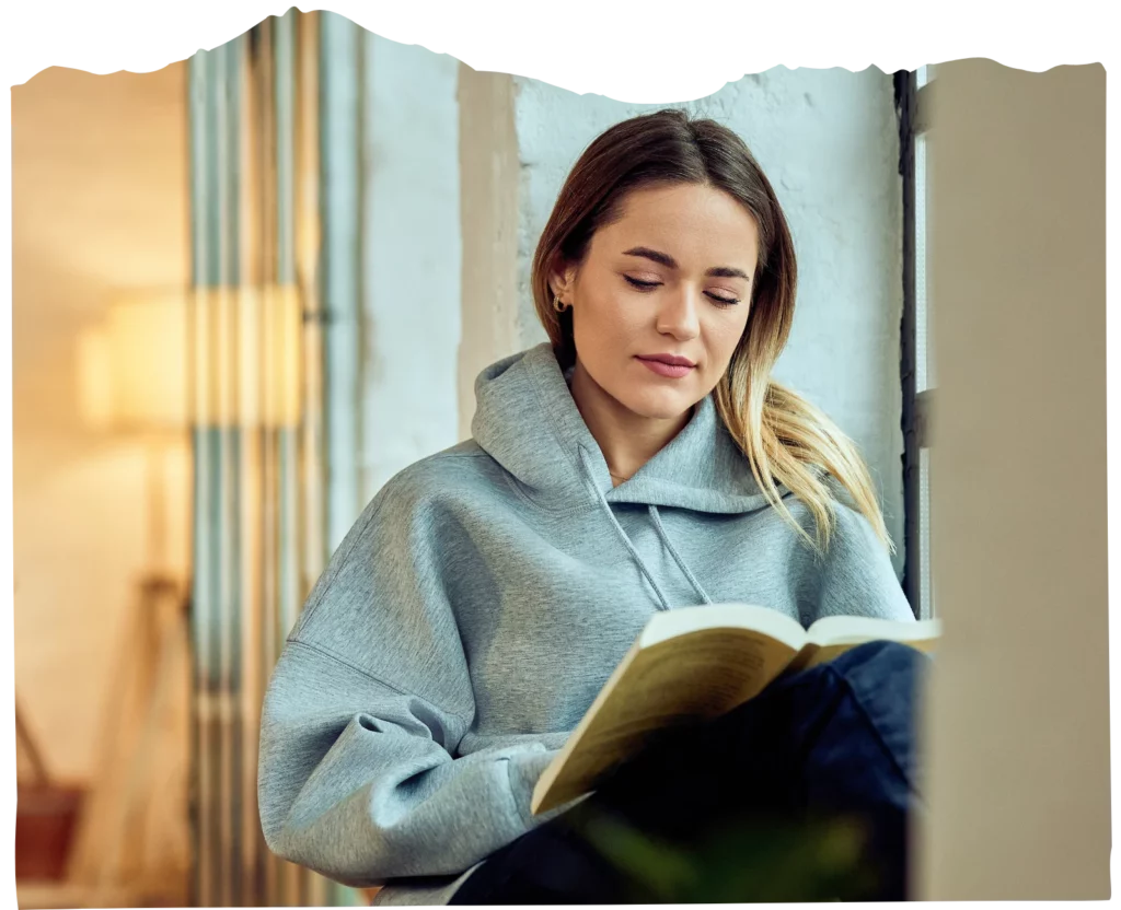 A woman in a gray hoodie sits by a window, reading a book with a calm expression, embracing a moment of continuing care for herself.