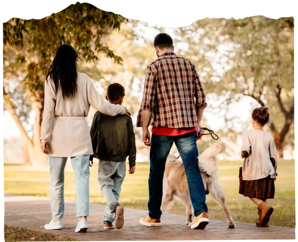 A family of four and a dog walk together on a paved path in a park, viewed from behind, surrounded by trees and greenery—a perfect scene for a Family Program outing.