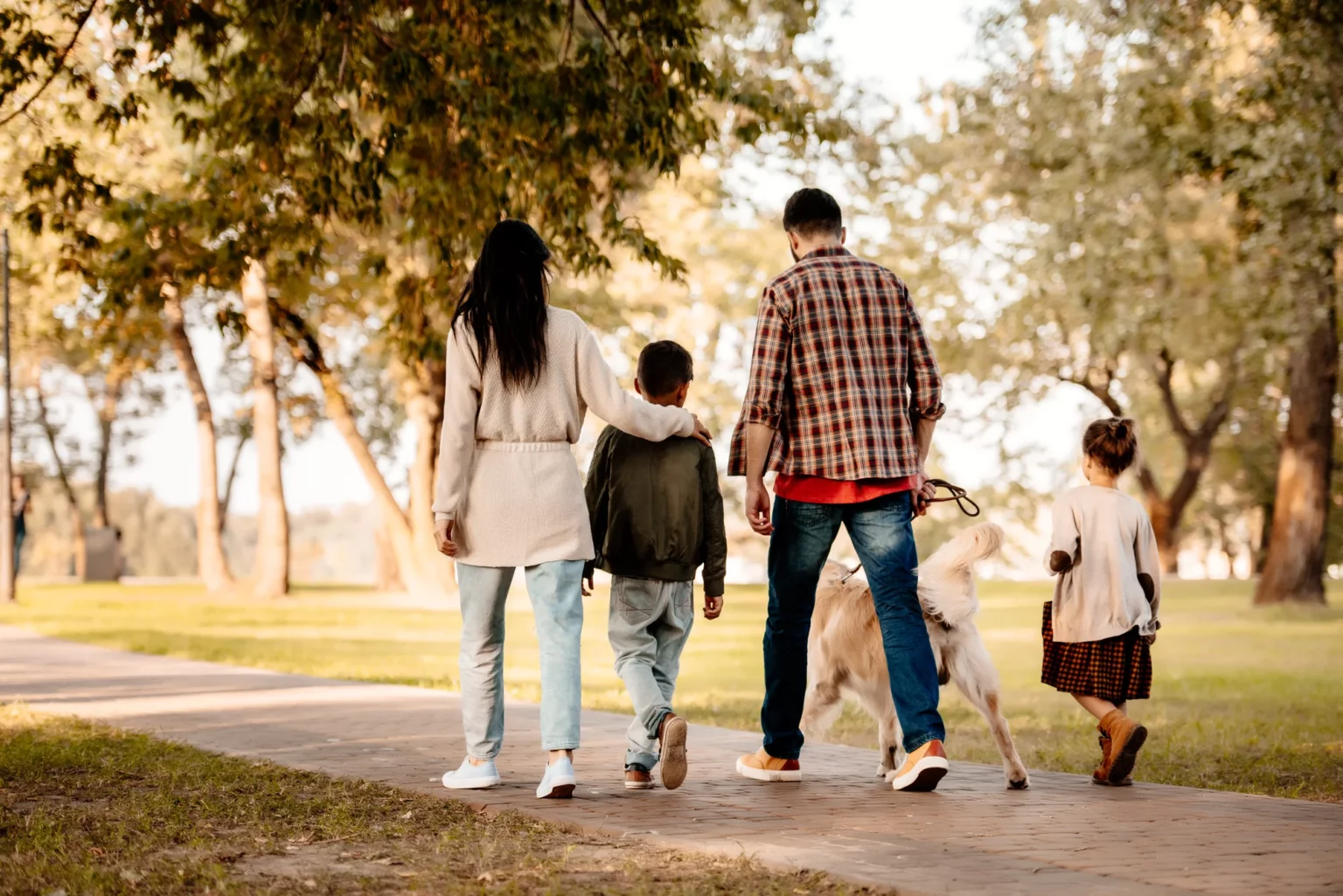 A family of four and a dog walk together on a paved path in a park, surrounded by trees and greenery near a local treatment center.