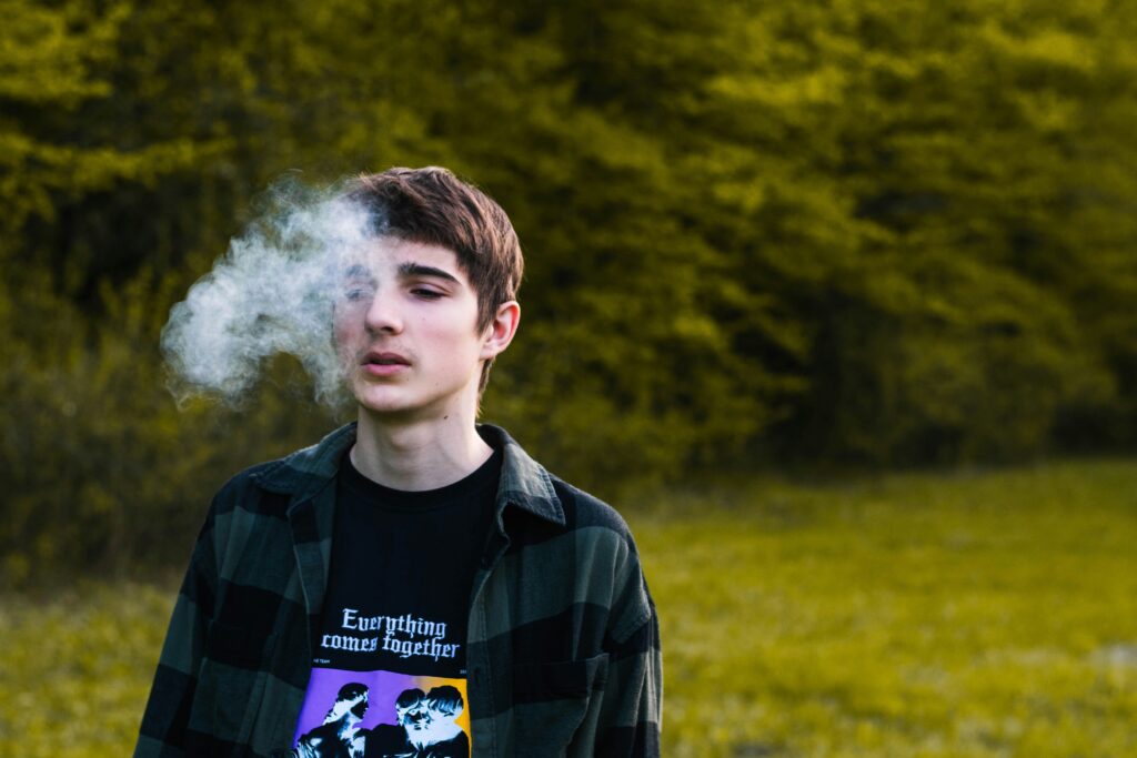 A young person stands in a grassy area with trees in the background, exhaling a cloud of smoke that partially covers their face—a subtle nod to substance abuse. They wear a dark flannel shirt over a graphic T-shirt.