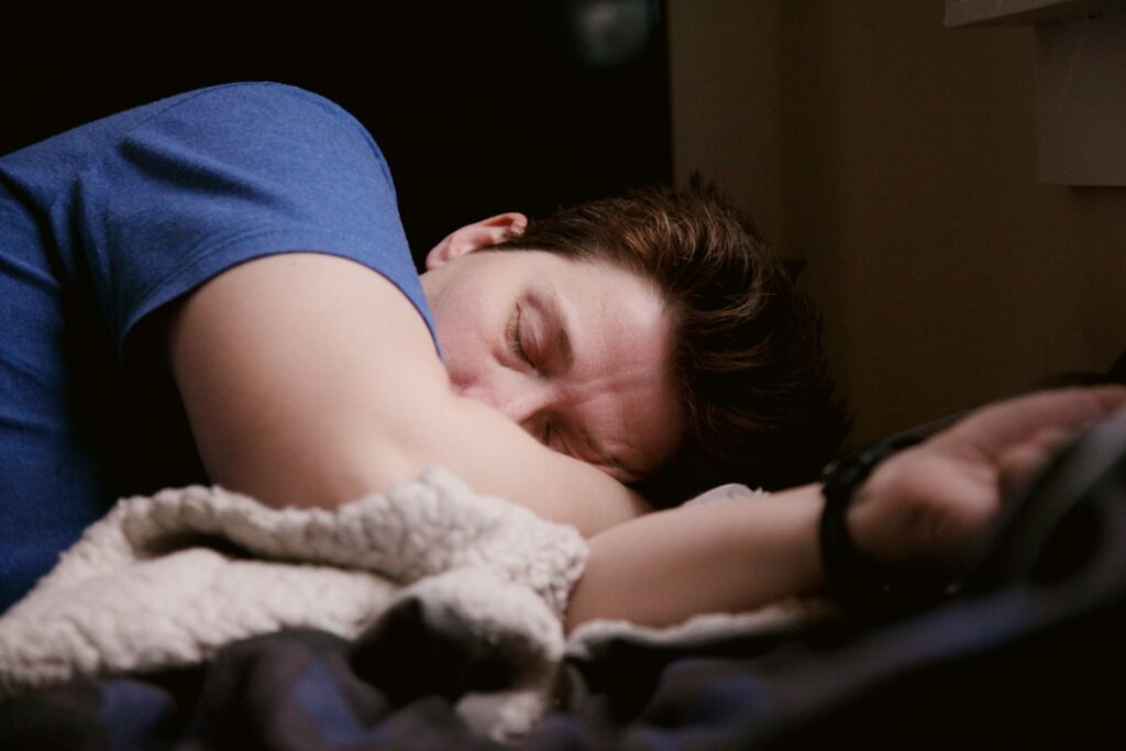 A person with short brown hair wearing a blue shirt is lying on their side, asleep, with their arm under their head and a soft blanket nearby—perhaps resting after a long day at outpatient rehab. The setting appears to be indoors.
