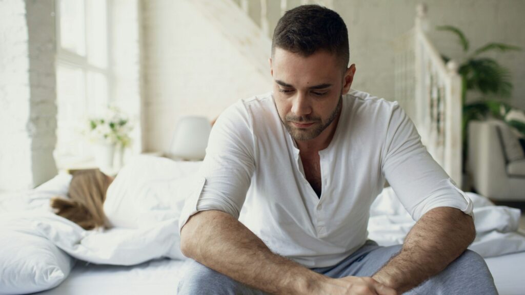 A man with short brown hair and a beard sits on a bed, looking down with his hands clasped. Another person is partially visible in the background, under the covers.