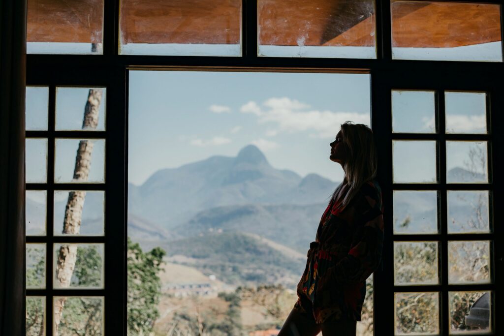 Silhouette of a woman in a colorful robe leaning against a window frame, looking out at a hazy mountain landscape.