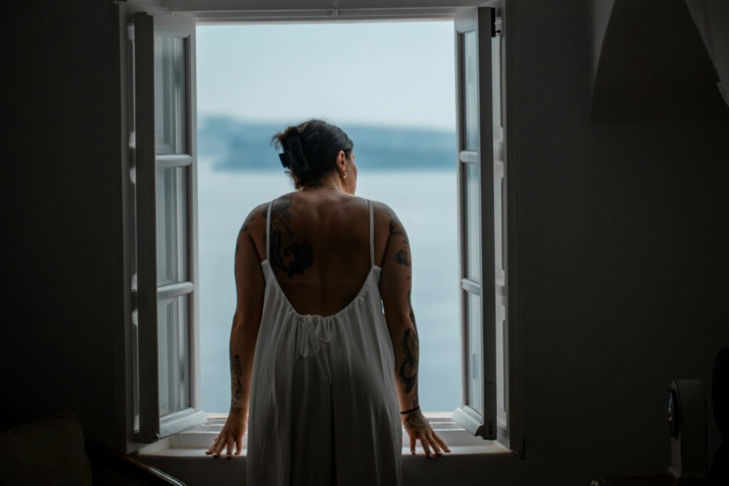 A woman with tattoos, wearing a white dress, stands at an open window looking out toward a blurred, serene body of water—finding peace after outpatient rehab. The scene is softly lit and tranquil.