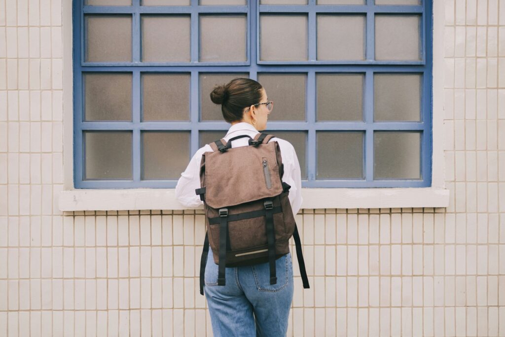A person with a bun hairstyle and glasses stands facing a frosted window with blue grid frames, wearing a brown backpack, white shirt, and blue jeans against a tiled wall, perhaps considering what to drink instead of alcohol.