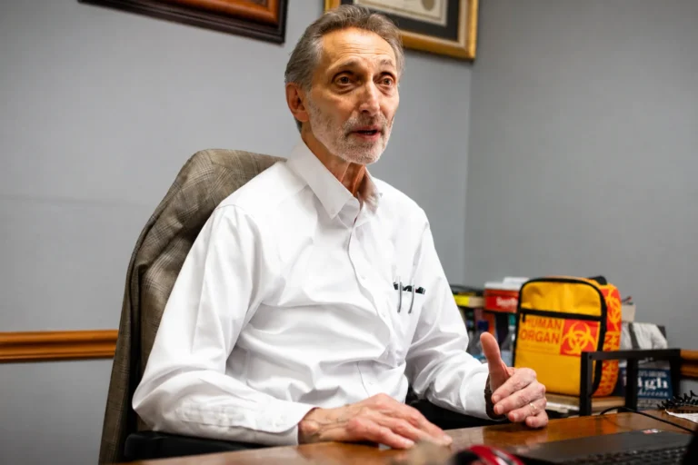 On older man in a white oxford shirt speaking while sitting at a desk