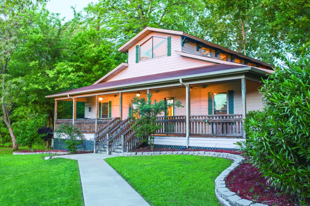 Palmetto two-story residential house with a large front porch, wooden railings, and green shutters, surrounded by trees and landscaped yard.